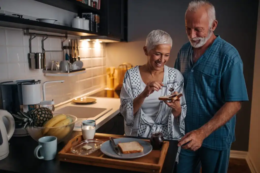 An older couple stands in a cozy kitchen, smiling as the woman spreads jam on toast. A breakfast tray with bread, milk, and fruit sits on the counter. The man stands close, watching her with affection.