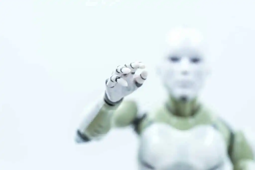 A humanoid robot with an outstretched hand is reaching forward. The image is softly focused, highlighting the robots white and metallic arm and hand against a bright, blurred background.