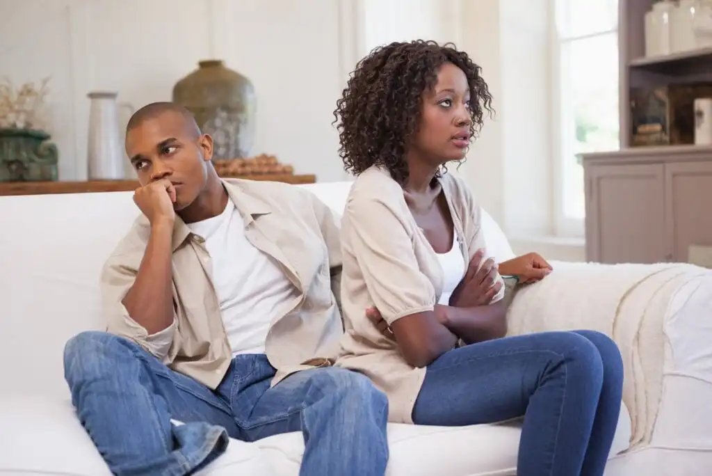 A young couple sits on a white couch looking upset. The man rests his chin on his hand, appearing frustrated, while the woman sits with her arms crossed, looking away. Both seem unhappy or in disagreement.