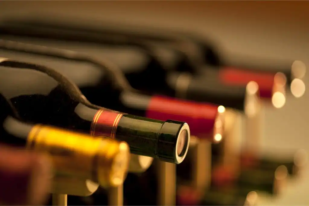 Close-up of several wine bottles lying on their sides in a wine rack, showing the necks and tops of the bottles with various colored foil caps. The background is softly blurred.