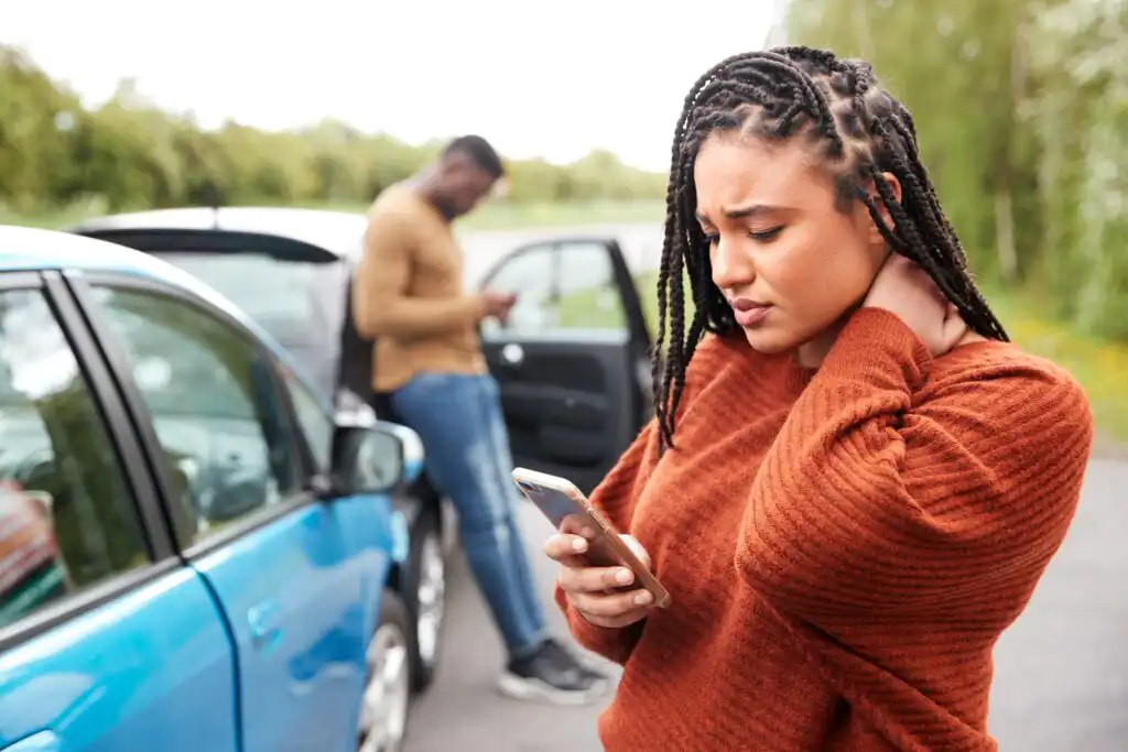 A woman stands by a blue car, looking at her phone with a worried expression and rubbing her neck. In the background, a man is on his phone near an open car door. Both cars seem to be stopped on the roadside.