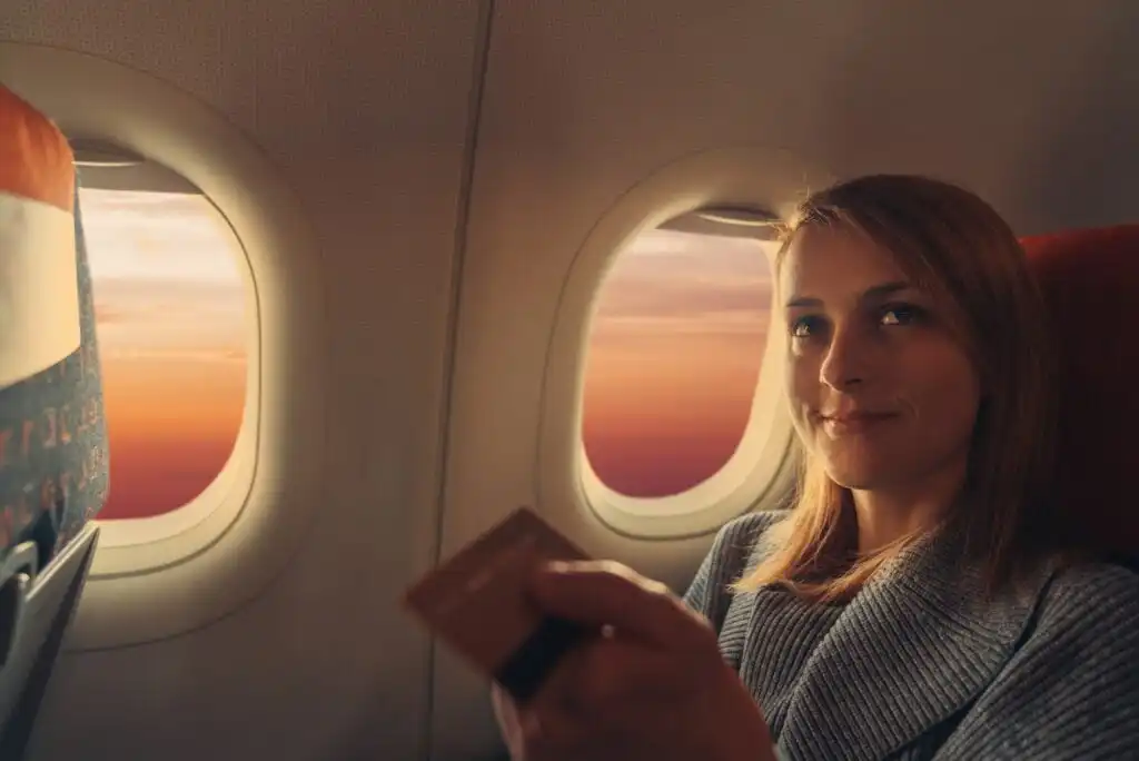 A woman sits by an airplane window at sunset, holding a credit card and smiling slightly, with warm sunlight illuminating the cabin.