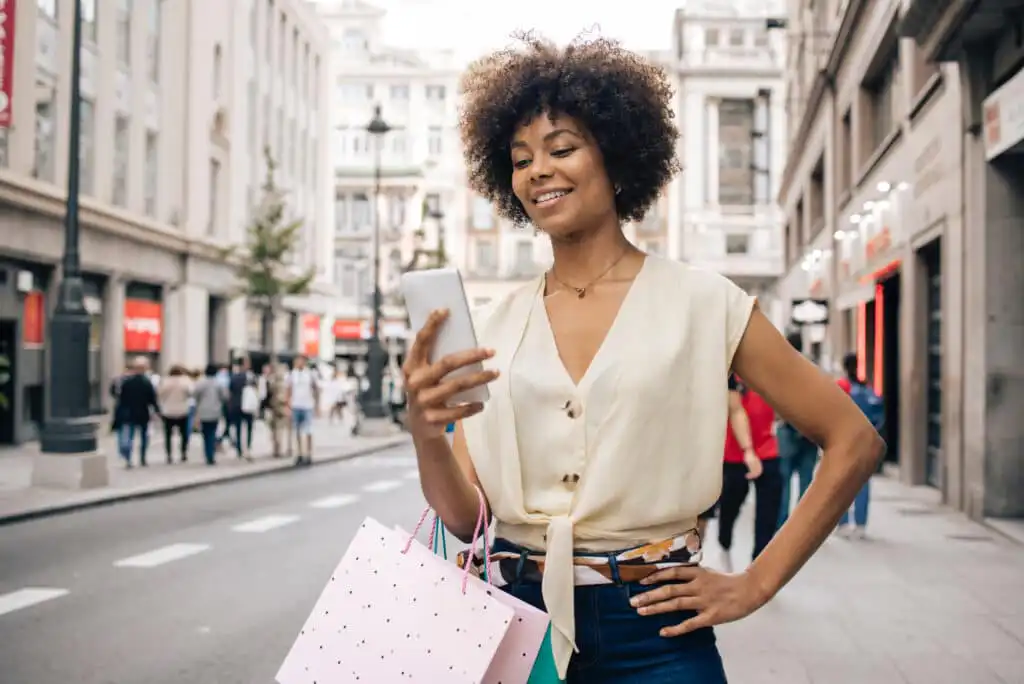 A woman stands on a city street holding shopping bags and smiling while looking at her smartphone, with buildings and people visible in the background.