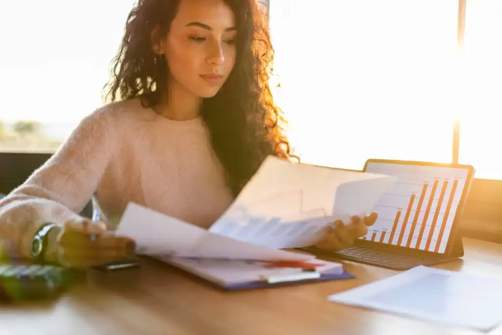 A woman with curly hair reviews documents with charts at a desk, with a tablet displaying a bar graph and sunlight streaming through a window in the background.