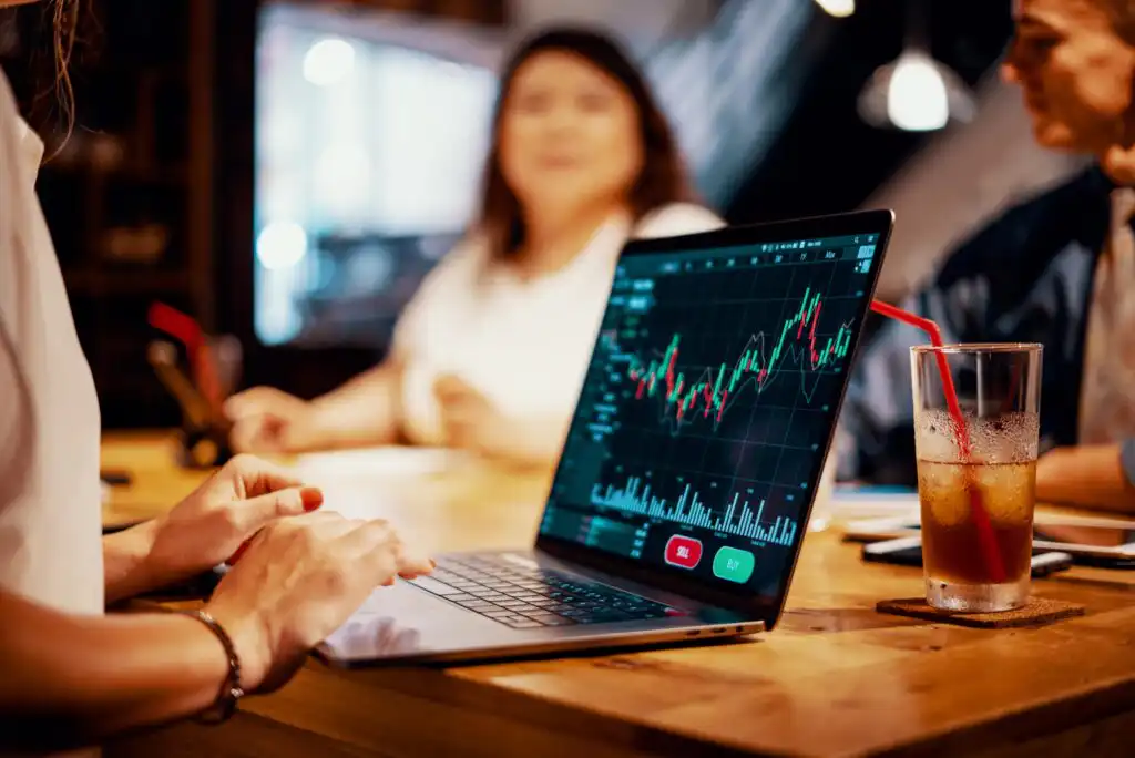 A person works on a laptop displaying a financial chart with red and green candlesticks at a wooden table. Two people sit in the background, and a drink with a red straw is on the table.