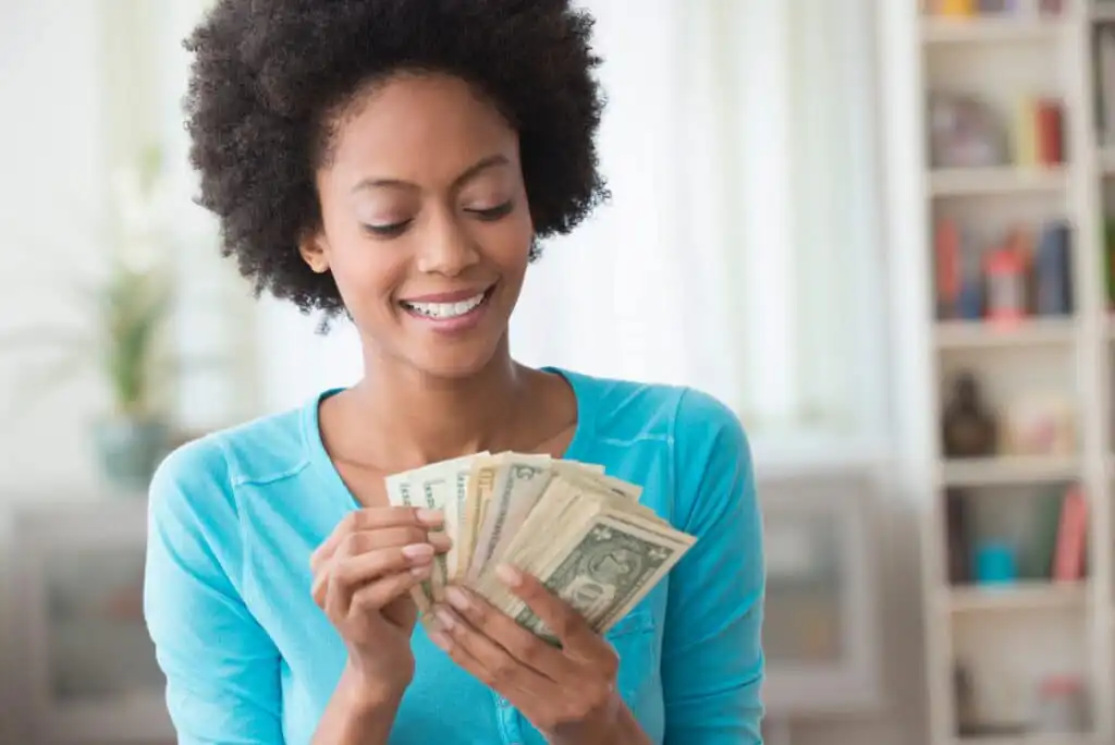 A smiling woman in a blue top counts a stack of U.S. dollar bills, standing indoors with soft light and shelves blurred in the background.