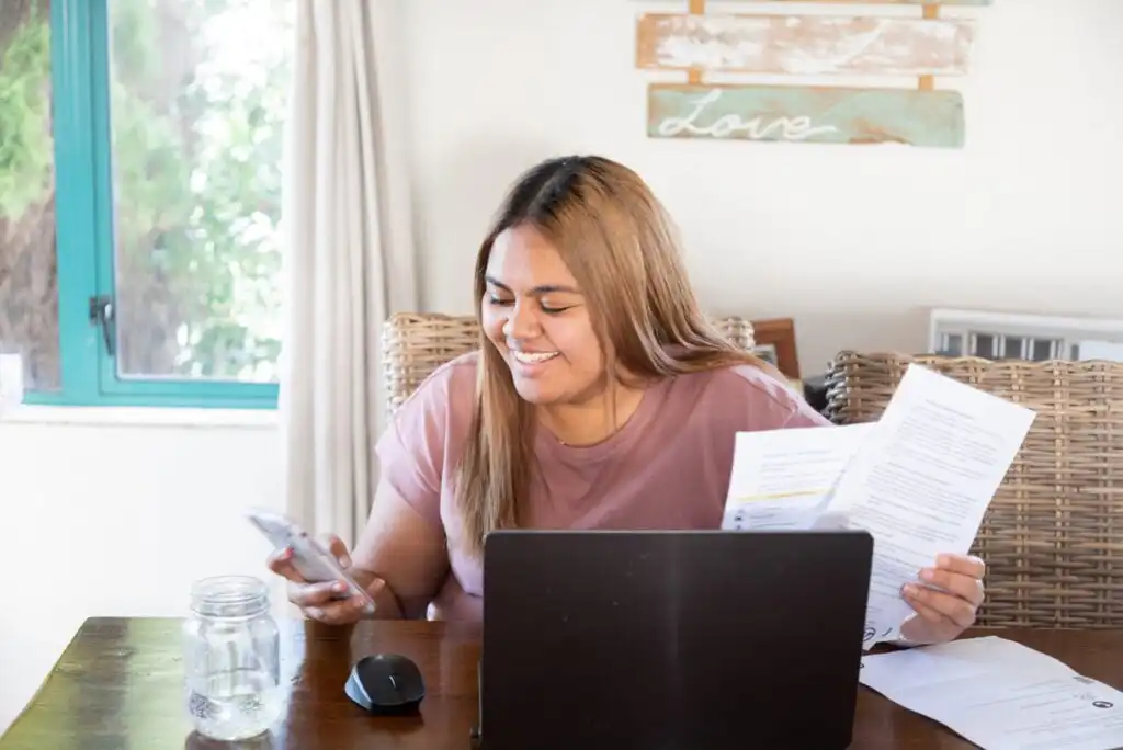 A woman sits at a table with a laptop, holding documents in one hand and looking at her phone, smiling. A glass jar, mouse, and “Love” sign are also visible on the table and wall behind her.