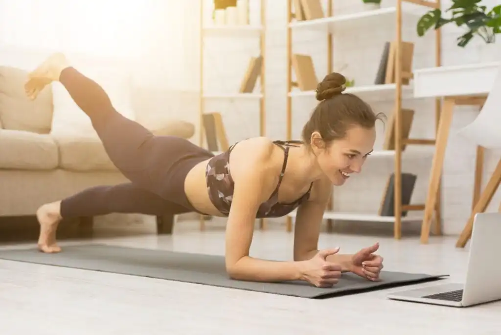 Young woman in workout clothes smiling while doing a plank leg raise on a yoga mat at home, watching a laptop, with sunlight streaming in and shelves in the background.