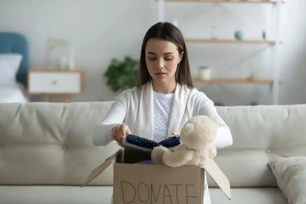 A woman sits on a couch placing clothes into a cardboard box labeled DONATE, with a teddy bear also visible in the box.