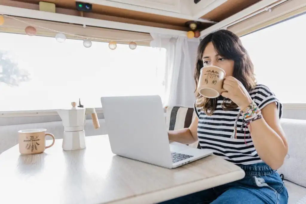 A woman sits at a table in a bright room, working on a laptop and drinking from a mug. She wears a striped shirt and beaded bracelets, with another mug and a coffee maker on the table nearby.