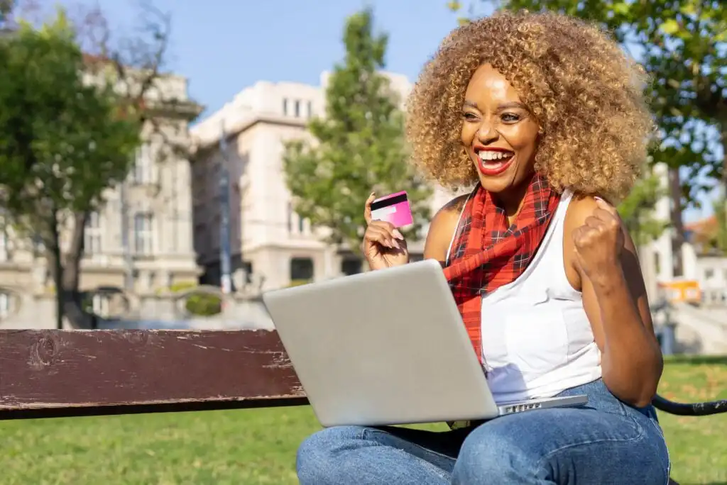 A smiling woman sitting on a bench outdoors holds a credit card and uses a laptop, looking excited and happy. Trees and buildings are visible in the background on a sunny day.