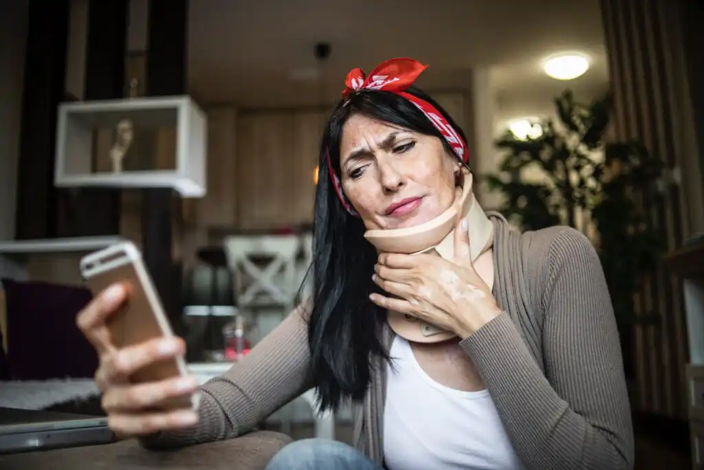 A woman wearing a neck brace and a red bandana sits indoors, looking concerned while holding a smartphone in one hand and touching her neck with the other.