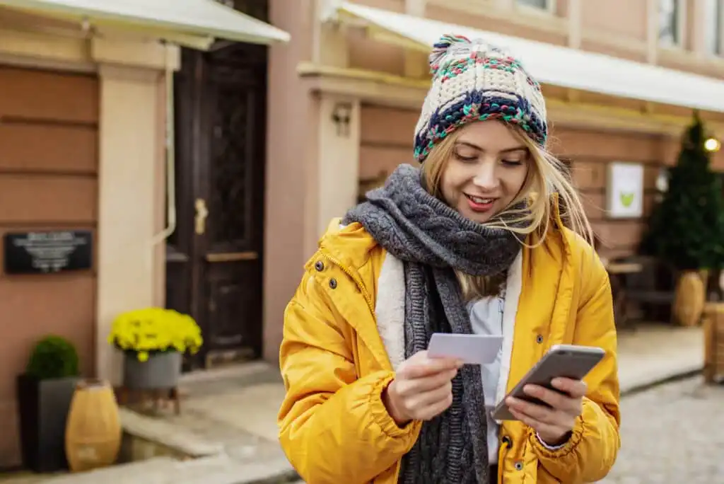A woman in a yellow jacket and colorful knit hat stands outside, holding a card in one hand and using a smartphone with the other. She is smiling and appears to be focused on the phone.