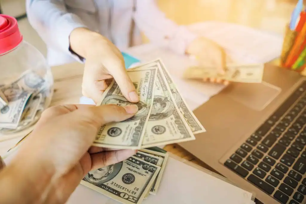 Close-up of two hands exchanging U.S. dollar bills near a laptop and a jar of money on a desk, suggesting a financial transaction or payment in a business or office setting.