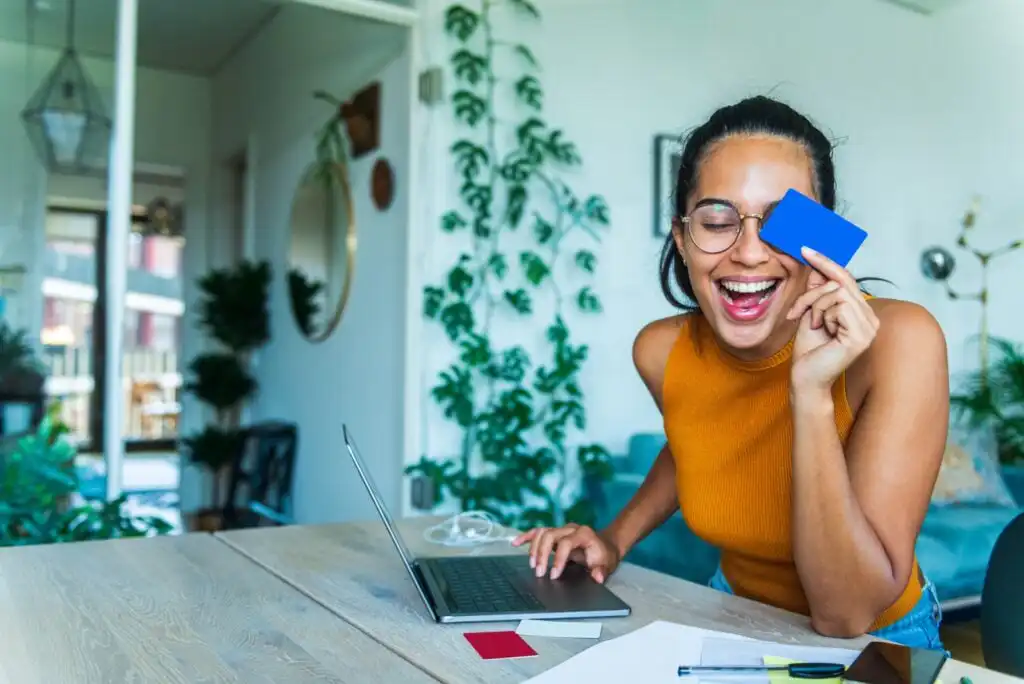 A smiling woman with glasses sits at a desk with a laptop, holding a blue card up to one eye. Papers, pens, and another card are on the tabletop. The background features green plants and a modern interior.