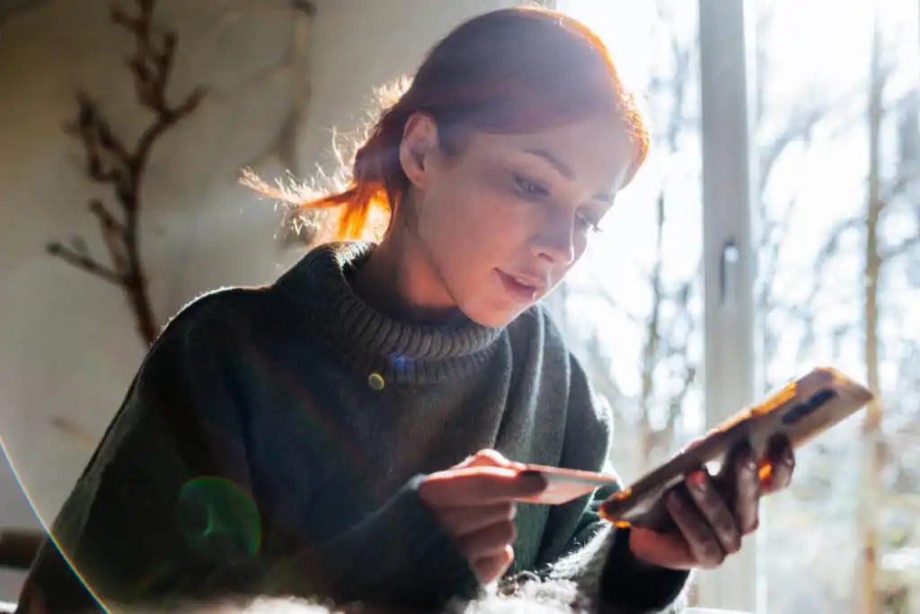 A young woman with red hair, wearing a dark sweater, sits indoors by a window while holding a smartphone in one hand and a credit card in the other, appearing focused on her online purchase. Sunlight streams in behind her.
