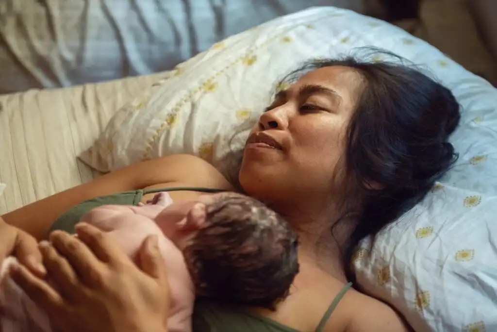 A woman lies on a bed, resting on a pillow with a floral pattern, gently holding a newborn baby against her chest. The scene appears calm and intimate, highlighting a post-birth moment.