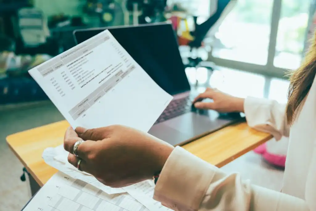 A person sitting at a desk holds a financial document while using a laptop. Other papers and receipts are spread out on the desk, indicating they are working on finances or budgeting.
