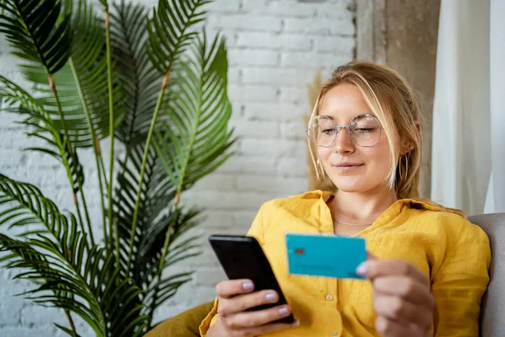 A woman wearing glasses and a yellow shirt sits indoors, holding a smartphone in one hand and a blue credit card in the other, with a large potted plant and a white brick wall in the background.