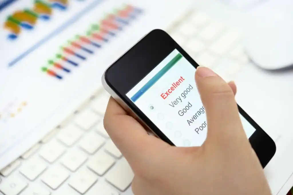 A person holds a smartphone displaying a survey with options: Excellent, Very good, Good, Average, Poor. In the background, a computer keyboard and blurred charts are visible.