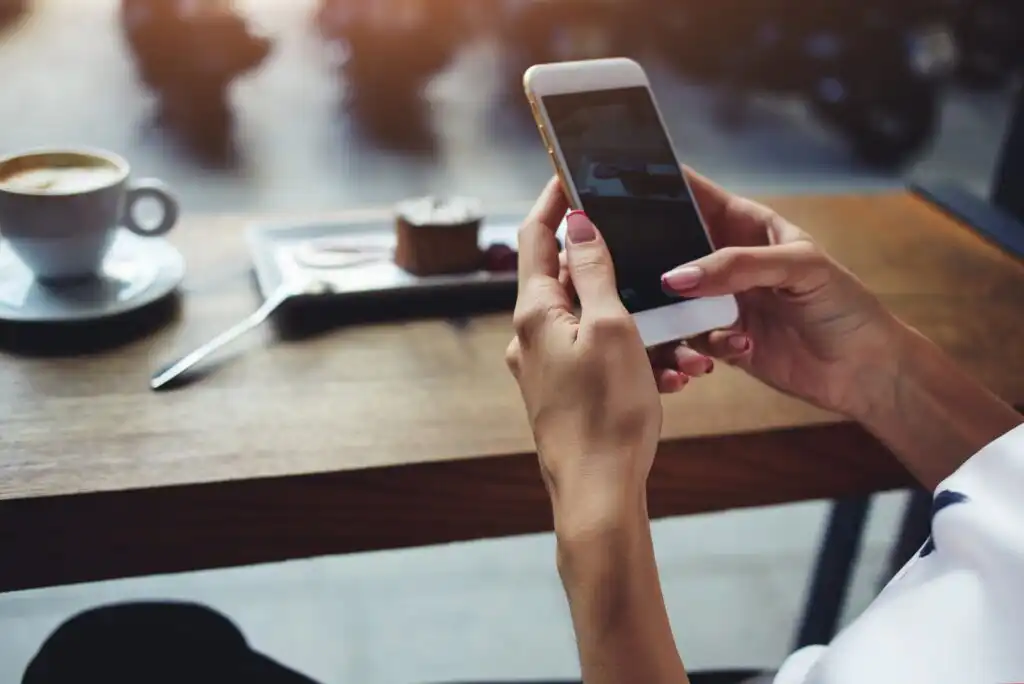 A person holding a smartphone at a wooden table, with a cup of coffee, a dessert, a fork, and a blurred background. The scene suggests a relaxed café setting.