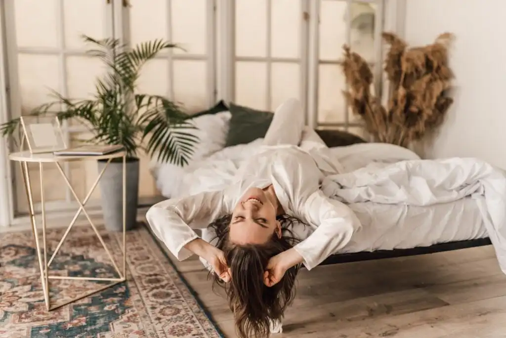 A woman in white pajamas lies playfully on a bed with her head hanging upside down, smiling and holding her hair. The room is bright with large windows, plants, and cozy decor.