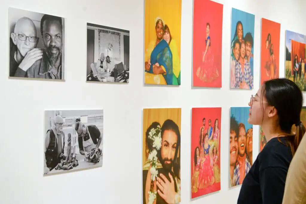 A woman stands in front of a gallery wall displaying colorful and black-and-white portrait photographs of diverse people and families, observing the artwork closely.