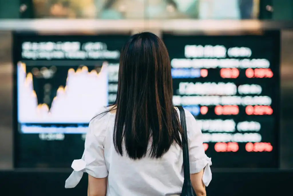 A woman with long dark hair, seen from behind, stands in front of large electronic screens displaying stock market charts and financial data.