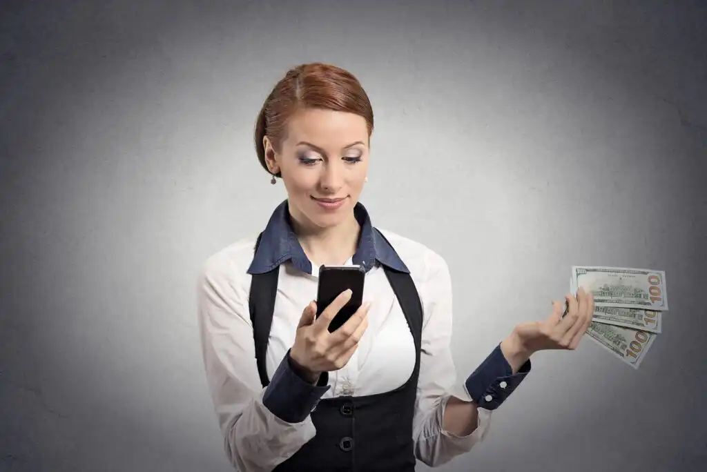 A woman in business attire smiles while looking at her smartphone, holding several US $100 bills in her other hand, against a plain gray background.