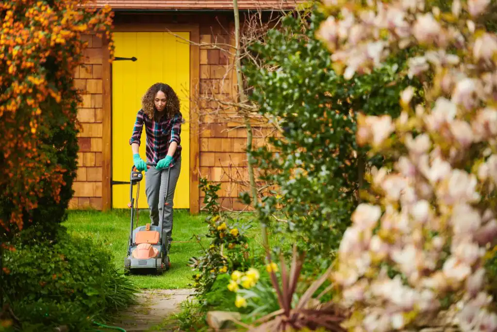 A person with curly hair and a plaid shirt mows the lawn in a garden, surrounded by blooming plants and greenery, with a bright yellow door on a wooden shed in the background.