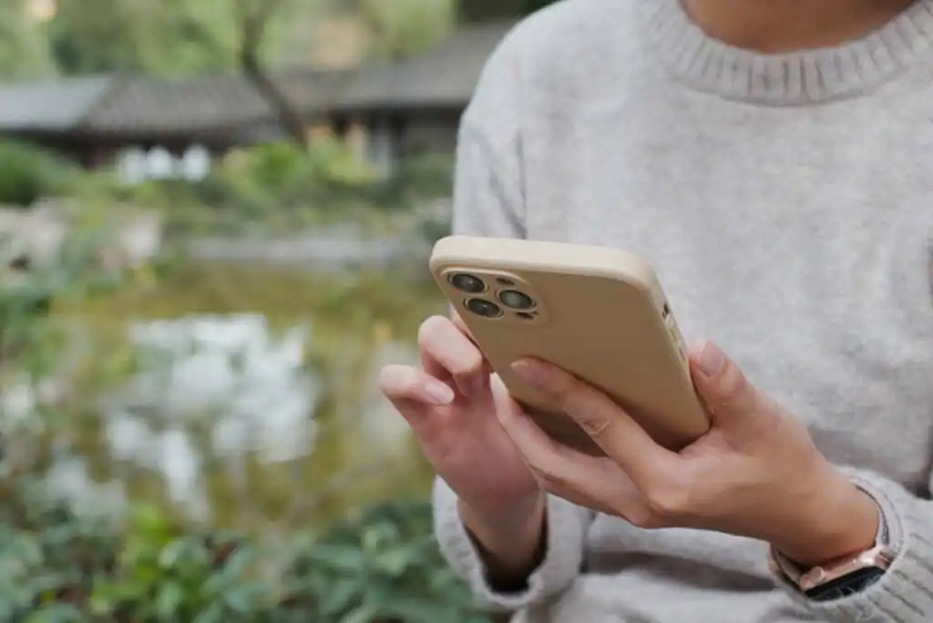 A person in a gray sweater holds a beige smartphone with both hands outdoors, using their thumb to interact with the screen. The blurred background shows greenery and a small pond.