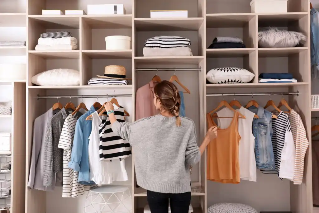 A woman stands in front of an organized wardrobe, looking at clothes on hangers. Shelves above hold folded garments, hats, and storage boxes, while assorted tops and jackets hang neatly below.