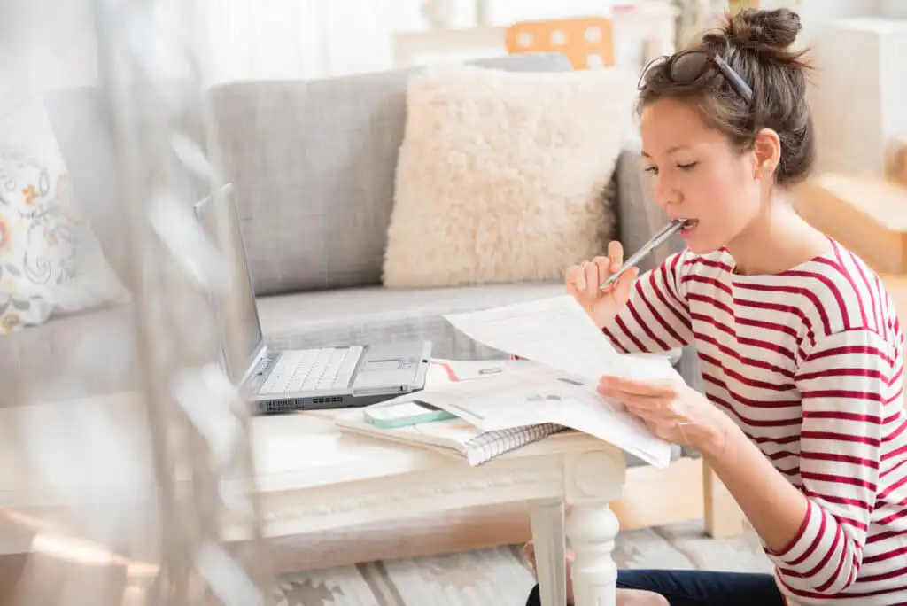 A woman in a striped shirt sits at a small table with a laptop and papers, thoughtfully holding a pen to her mouth while reviewing documents in a cozy living room.