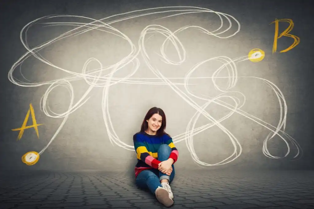 A young woman sits on the floor smiling, with a complex, tangled white line drawn on the wall behind her, connecting two yellow points labeled A and B.
