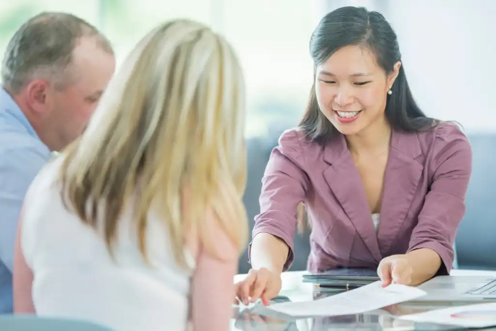 A woman in a mauve blazer smiles while handing a document to a blonde woman across the table. A man sits beside them. They appear to be in a business meeting or consultation in a bright office setting.