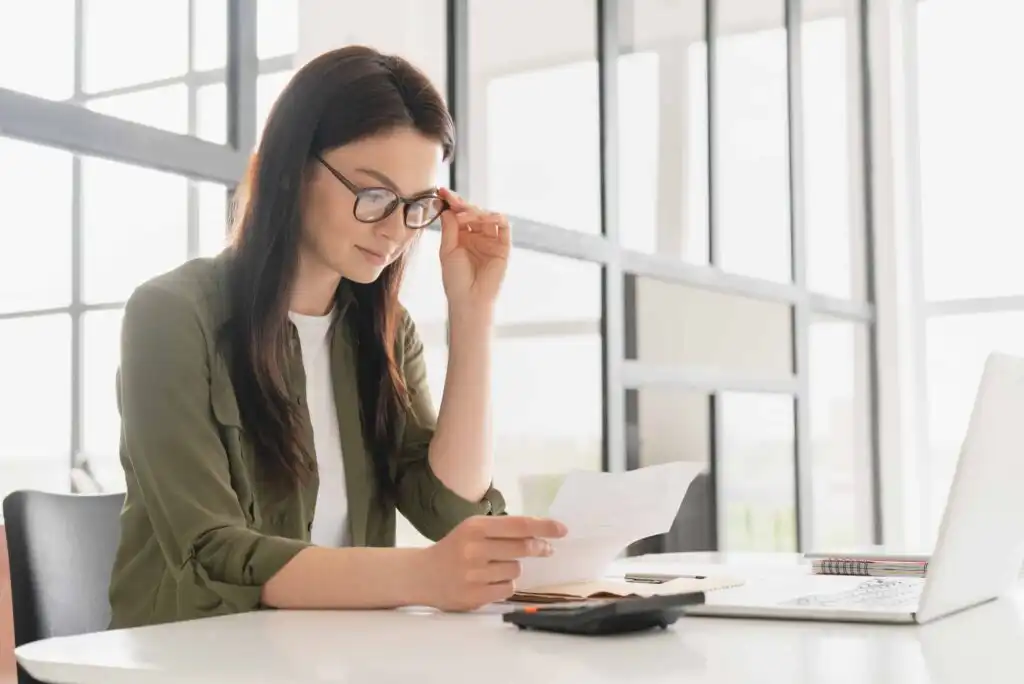 A woman wearing glasses sits at a desk, holding a piece of paper and looking at it thoughtfully. A calculator, notebook, and laptop are on the table in a bright, modern office space.