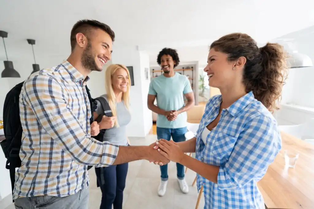 Four young adults smile in a brightly lit room; two people in the foreground shake hands warmly, while the other two stand in the background, watching and smiling.