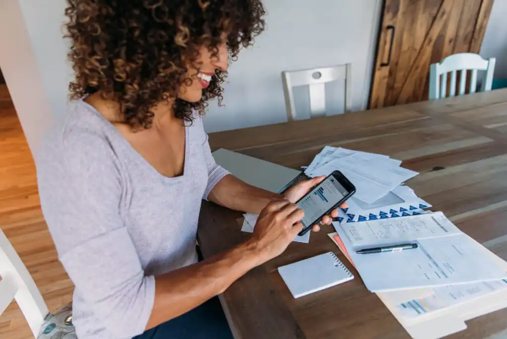 A woman sits at a table smiling while using a smartphone. Papers, a notepad, pen, and envelopes are spread across the table, suggesting she is managing finances or paying bills.