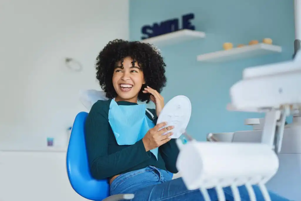 A young woman with curly hair smiles while sitting in a dental chair, holding a handheld mirror. She wears a dental bib and appears happy in a modern, brightly lit dental office.