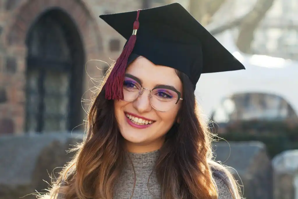 A young woman wearing glasses and a graduation cap with a maroon tassel smiles outdoors, with blurred stone buildings and trees in the background.