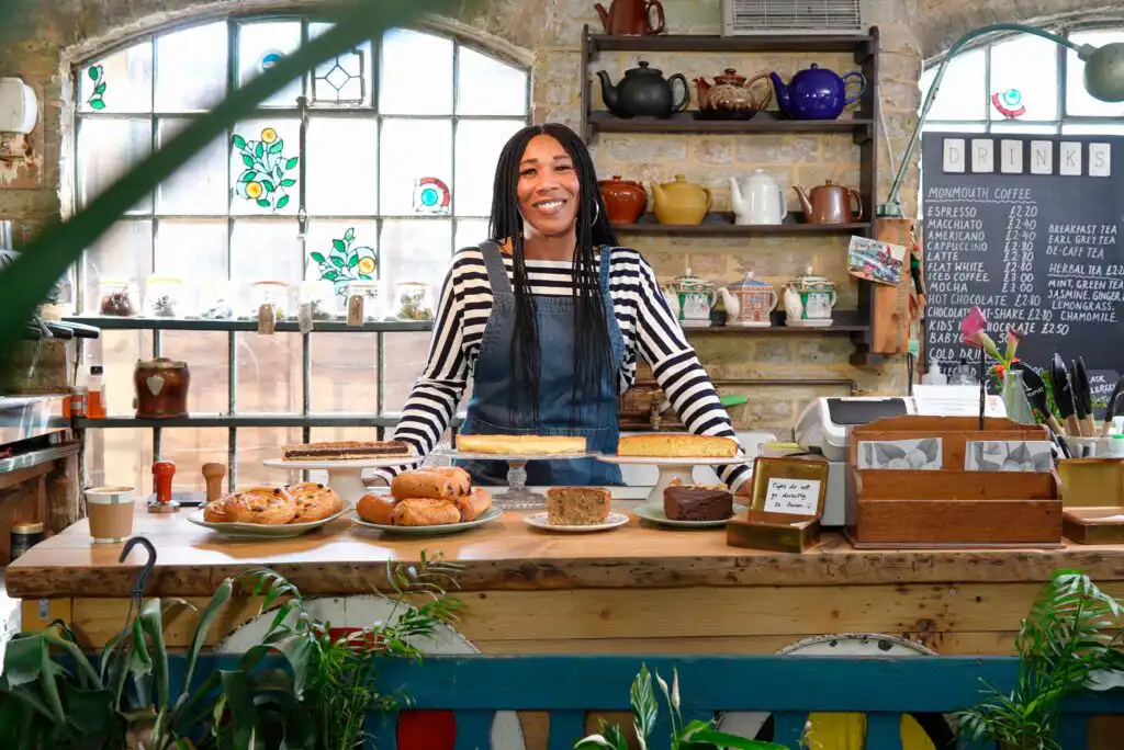 A smiling woman stands behind the counter of a cozy café, displaying various pastries and cakes. Behind her are shelves with teapots, cups, and a chalkboard menu. Green plants frame the warm, inviting scene.