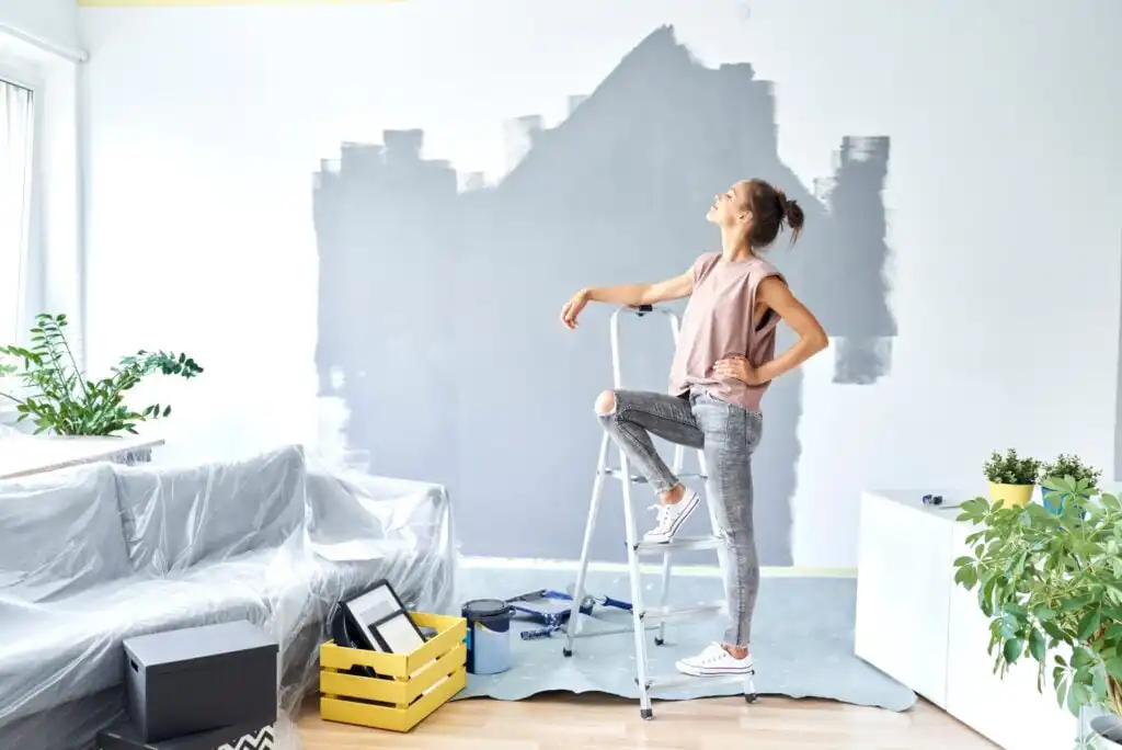 A woman stands on a stepladder, looking up at a partially painted gray wall in a bright living room. Furniture is covered in plastic; painting supplies and plants are visible around the room.