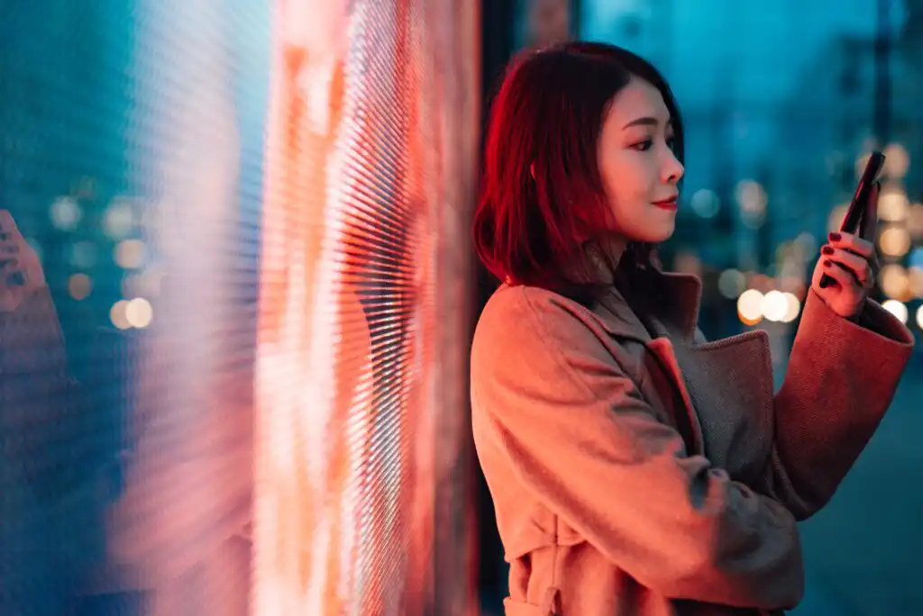 A woman in a tan coat stands against a brightly lit wall at night, looking at her smartphone with a slight smile. City lights blur in the background, creating a colorful, urban atmosphere.