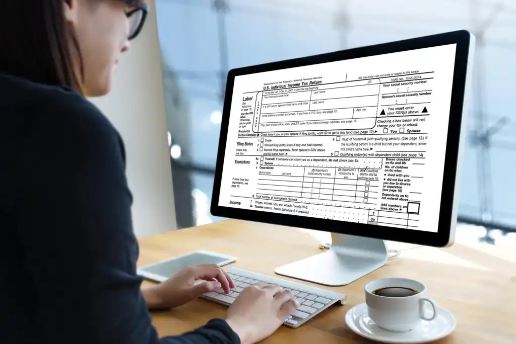 A person sits at a desk using a computer, with a U.S. tax form displayed on the monitor. The individual is typing on a keyboard, and the workspace includes a coffee cup and a phone.