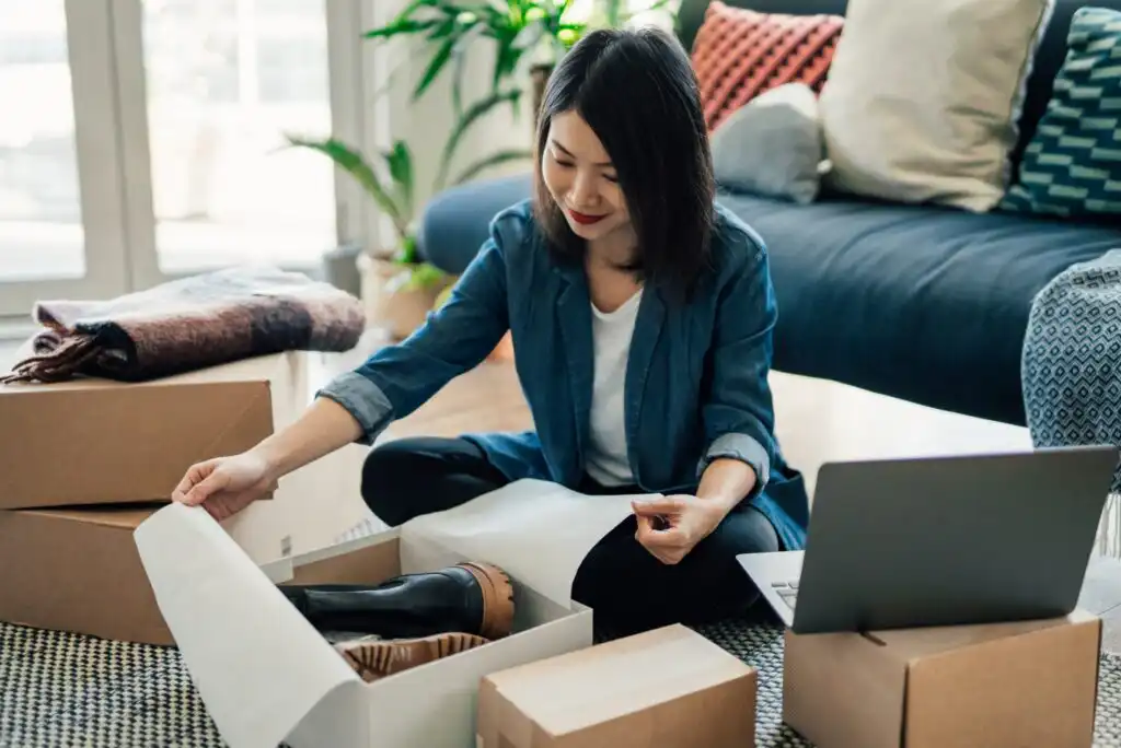 A woman sits on the floor in a living room, smiling as she unboxes a pair of black boots. She is surrounded by cardboard boxes and has an open laptop nearby. Cozy decor and plants are visible in the background.