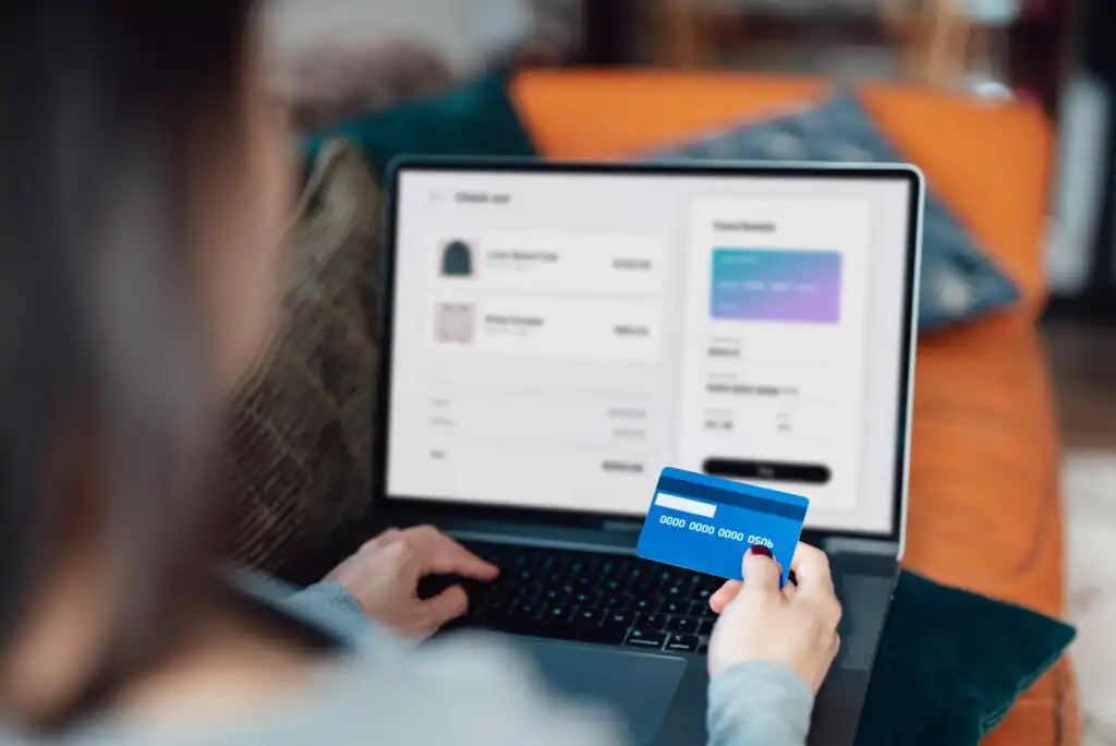 Person holding a blue credit card while making an online purchase on a laptop; the screen shows a checkout page with product details and payment information.