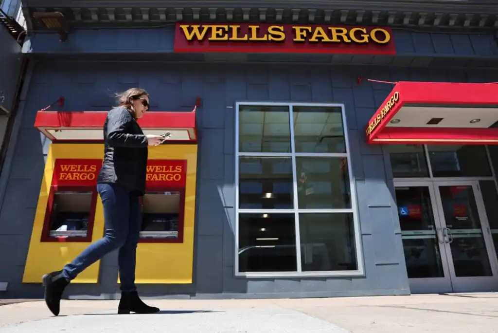 A person walks past a Wells Fargo bank branch with yellow and red signage, next to an ATM and large windows during the day.