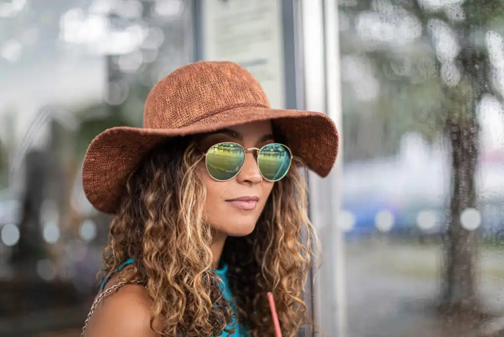 A woman with curly hair wears a wide-brimmed brown hat and round sunglasses, standing near a window on a rainy day, holding a drink with a pink straw.