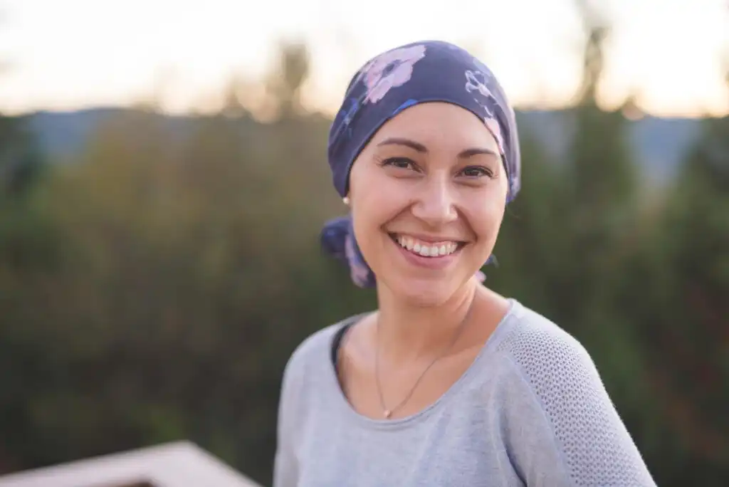 A woman wearing a floral headscarf and a grey top smiles warmly at the camera while standing outdoors, with blurred greenery in the background.