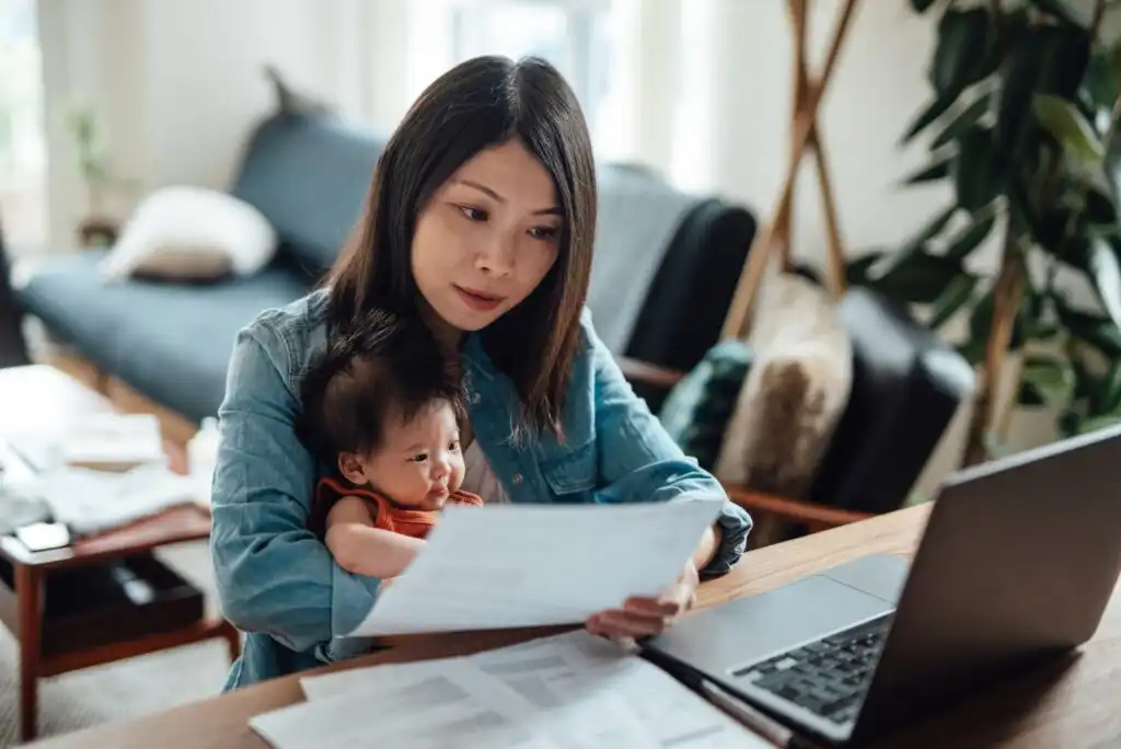 A woman sits at a desk holding a baby, looking at documents in her hand with an open laptop in front of her. The room has a couch, plants, and natural light from a window.